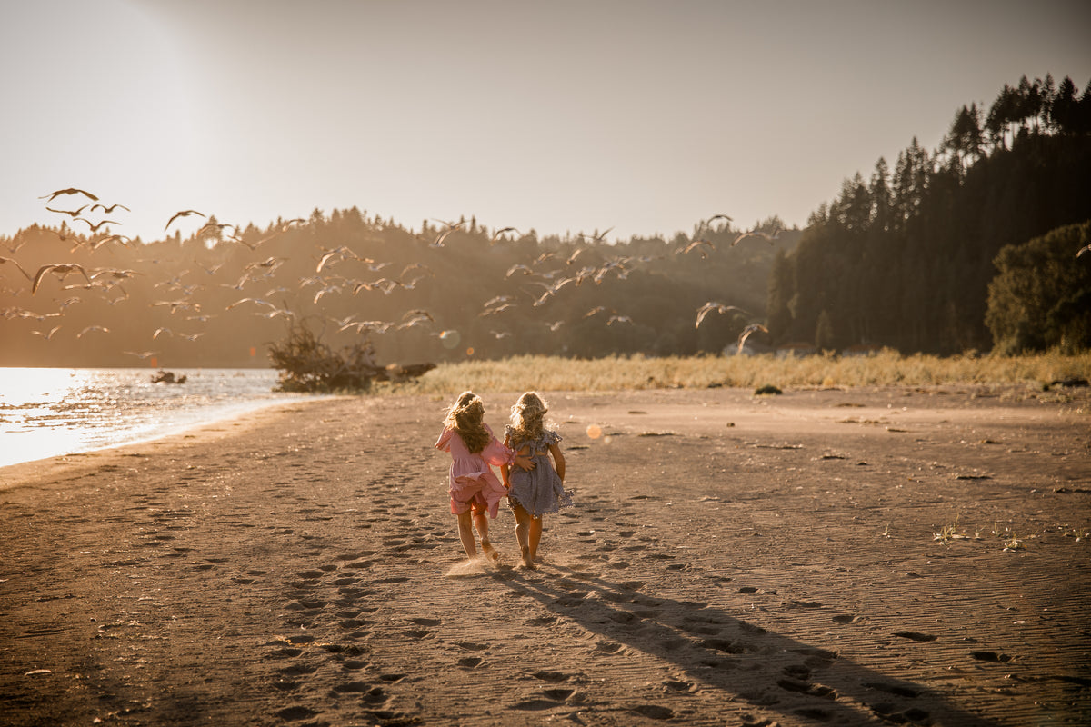 Two young girls running on a sandy beach at golden hour, with water, trees, and flying birds in the background.