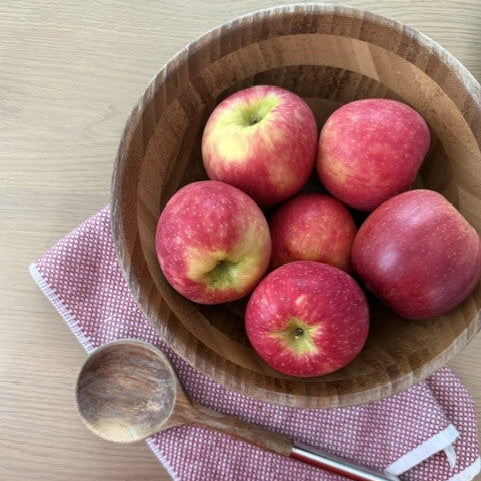 A wooden bowl filled with red apples on a light wood table, resting on a pink-and-white checkered cloth with a wooden spoon.