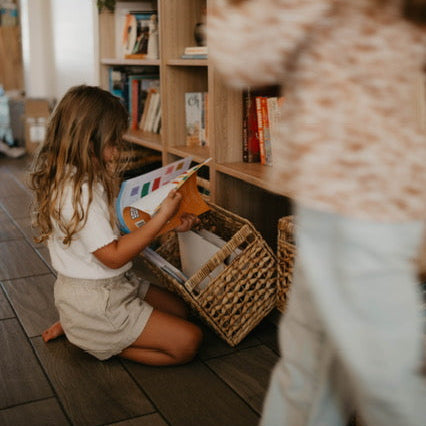 A young girl with long light brown hair sits on a dark wood floor in front of a tall wooden bookshelf, holding a colorful book. Another child in a beige patterned top and white pants is blurred in the foreground, reaching toward the bookshelf.