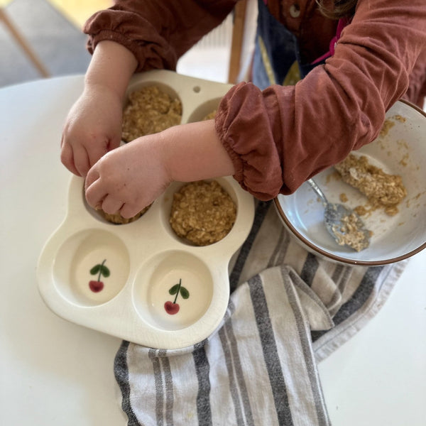 A close-up of a child's hands filling small compartments of a white silicone baby feeding tray with oatmeal. The tray features two cherry illustrations in the bottom compartments.