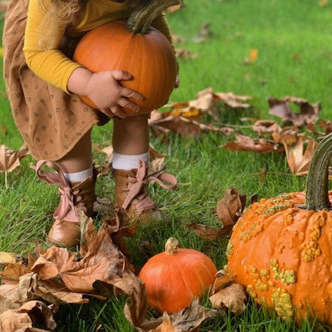 A child in a yellow top and brown dress holds an orange pumpkin on green grass surrounded by fallen leaves, with two pumpkins in the foreground.